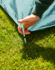 Hand holding light blue tent stake at teal Socialite Picnic Set base on grass